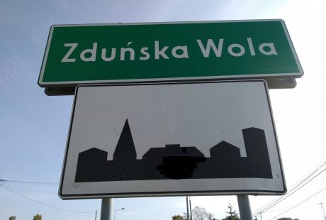 Na zdjęciu jest zielona tablica z nazwą miasta Zduńska Wola, pod którą wisi tablica biała oznaczająca teren zabudowany.