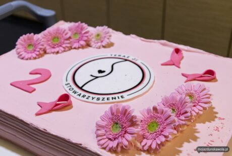 Duży różowy tort ozdobiony logo „taras uefa”, różowymi kwiatami gerbery i wstążkami, wystawiony na stole podczas imprezy.