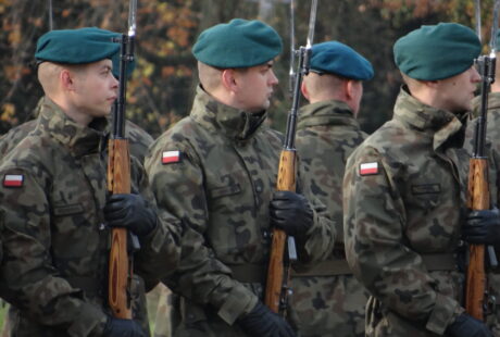 Czterech żołnierzy w mundurach kamuflażowych i zielonych beretach trzymających karabiny, stojących w kolejce podczas ceremonii wojskowej. flagi widoczne na ich ramionach.