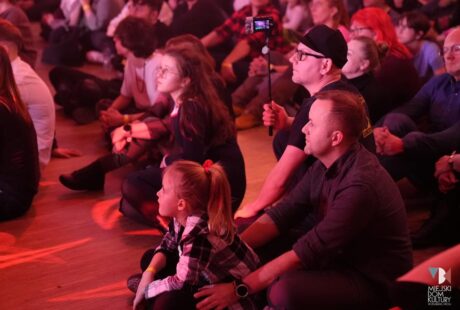 Zróżnicowana publiczność siedząca na podłodze w sali koncertowej i uważnie obserwująca występ, niektórzy rejestrowali ten moment smartfonami.