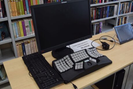 Komputerowa stacja robocza z monitorem Lenovo, dwiema klawiaturami i ergonomiczną podpórką pod nadgarstki na biurku w bibliotece.