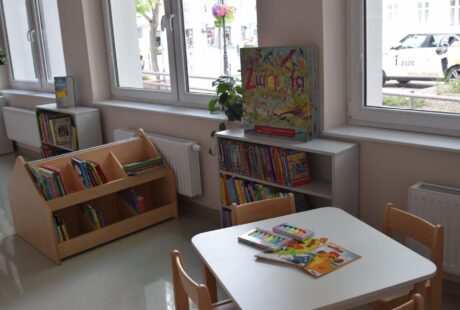 Kącik biblioteczny dla dzieci z półkami na książki, stołem z książkami i rośliną przy oknie, z widokiem na ulicę.