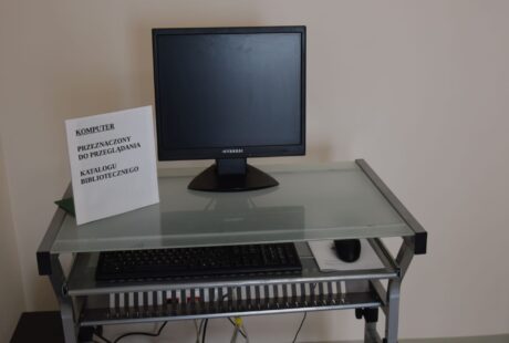 Konfiguracja komputera na szklanym biurku z monitorem, klawiaturą i myszą, oznaczona etykietą umożliwiającą przeglądanie katalogu w bibliotece.
