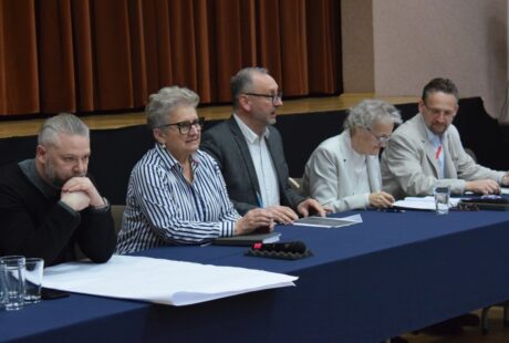 Pięciu członków panelu, trzech mężczyzn i dwie kobiety, siedzących przy stole z mikrofonami i dokumentami podczas wydarzenia odbywającego się w pomieszczeniu.