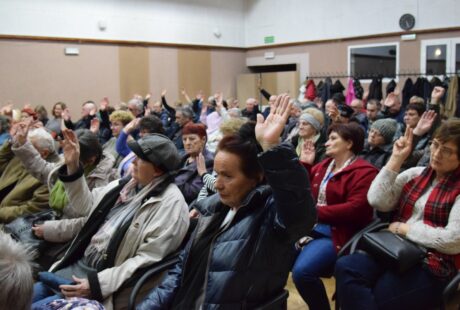 Grupa starszych osób podnoszących ręce w sali konferencyjnej, biorących udział w głosowaniu lub odpowiadaniu na pytania.