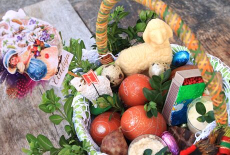 Kosz wielkanocny wypełniony dekorowanymi jajkami, świecą w kształcie baranka i drobnymi upominkami na drewnianym stole.