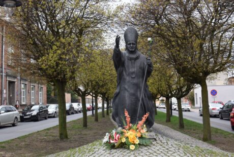 Pomnik postaci w szatach z brodą na wysadzanej drzewami ulicy, z kwiatowymi hołdami umieszczonymi u podstawy, w tle zaparkowanymi samochodami i budynkami.