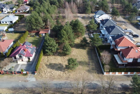 Widok z lotu ptaka na podmiejską dzielnicę z rzędami domów, niektóre z czerwonymi dachami, otoczoną drzewami i drogami.
