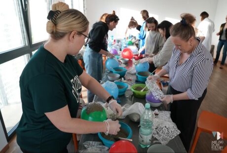 Dwie kobiety pracują razem nad projektem rękodzielniczym z użyciem pasty i balonów przy stole otoczonym innymi uczestnikami.