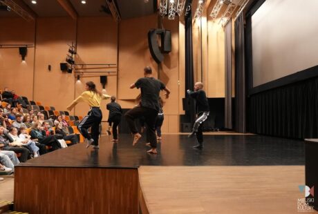 Czterech tancerzy występuje na scenie przed publicznością osadzoną w nowoczesnym teatrze.