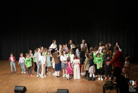 Grupa dzieci stojących na scenie z dorosłymi, współdziałających w ramach wydarzenia kulturalnego lub szkolnego, z publicznością na pierwszym planie.