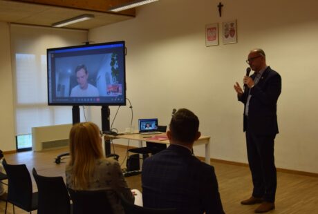 Mężczyzna wygłasza przemówienie w sali konferencyjnej, podczas gdy na ekranie wyświetlany jest wirtualny uczestnik z obecną publicznością.