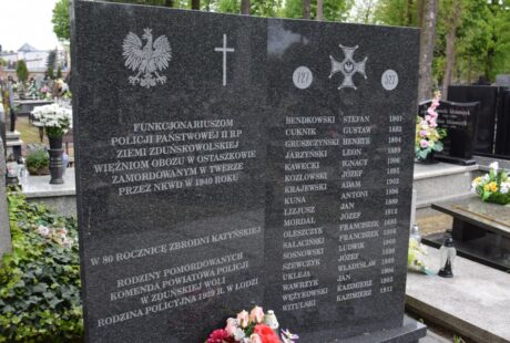 Ponury czarny kamień pamiątkowy ozdobiony polskimi symbolami i napisami, upamiętniający poległych funkcjonariuszy policji, z kwiatowymi hołdami u podstawy.