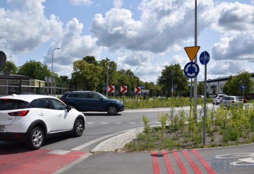 Kilka samochodów porusza się po rondzie w częściowo pochmurny dzień, z widocznymi znakami drogowymi, zielenią i przejściem dla pieszych.