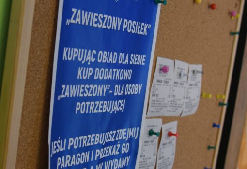 Polski napis na tablicy korkowej wyjaśnia zawieszony program posiłków, zachęcając ludzi do kupowania dodatkowych posiłków dla potrzebujących; paragony są przypięte w pobliżu.