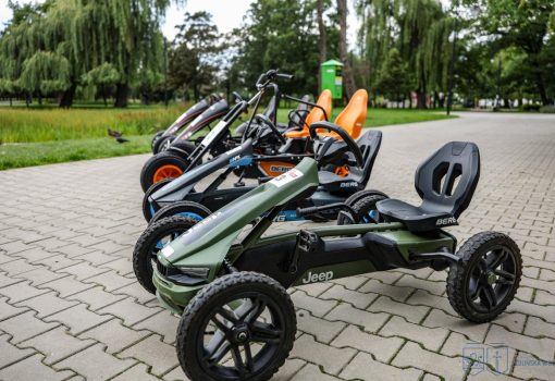 Kilka gokartów na pedały, w tym zielony model Jeepa, jest zaparkowanych na utwardzonej ścieżce w parku z drzewami i trawą w tle.
