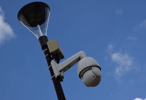 Słup latarni ulicznej z zamontowaną kamerą bezpieczeństwa i małą skrzynką kontrolną na tle błękitnego nieba z kilkoma chmurami.