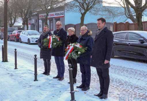 Pięć osób stoi na zaśnieżonej ulicy, trzymając wieńce z biało-czerwonymi wstążkami, pozując do zdjęcia przed zaparkowanymi samochodami i budynkiem.